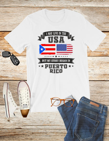 A white T-shirt reads, "I MAY LIVE IN THE USA BUT MY STORY BEGAN IN PUERTO RICO," with U.S. and Puerto Rican flags. Styled with jeans, white sneakers, glasses, and car keys on a wooden background.